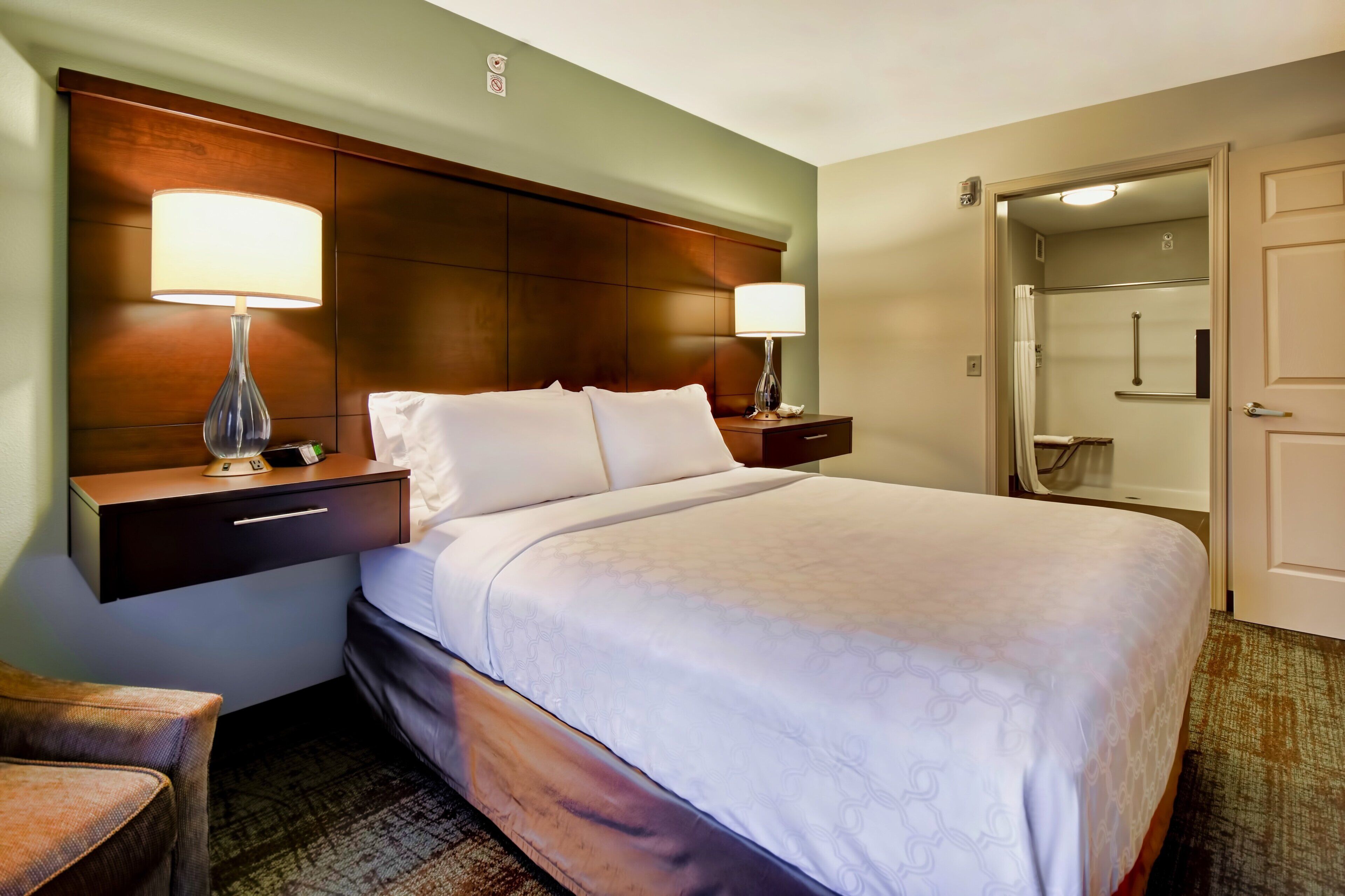 A hotel room with a bed, bedside lamps, a nightstand, a chair, and a shower area.