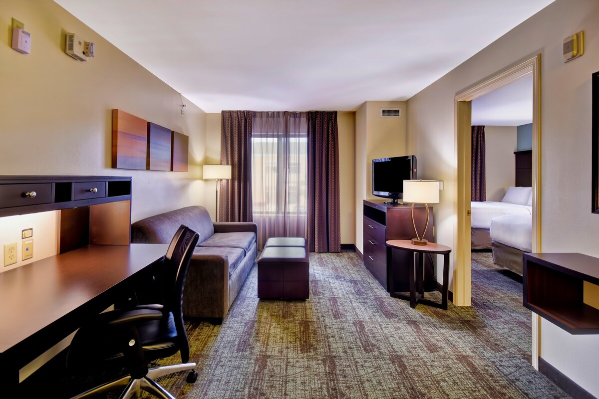A hotel room with a desk, chair, sofa, ottoman, TV, and a bed.