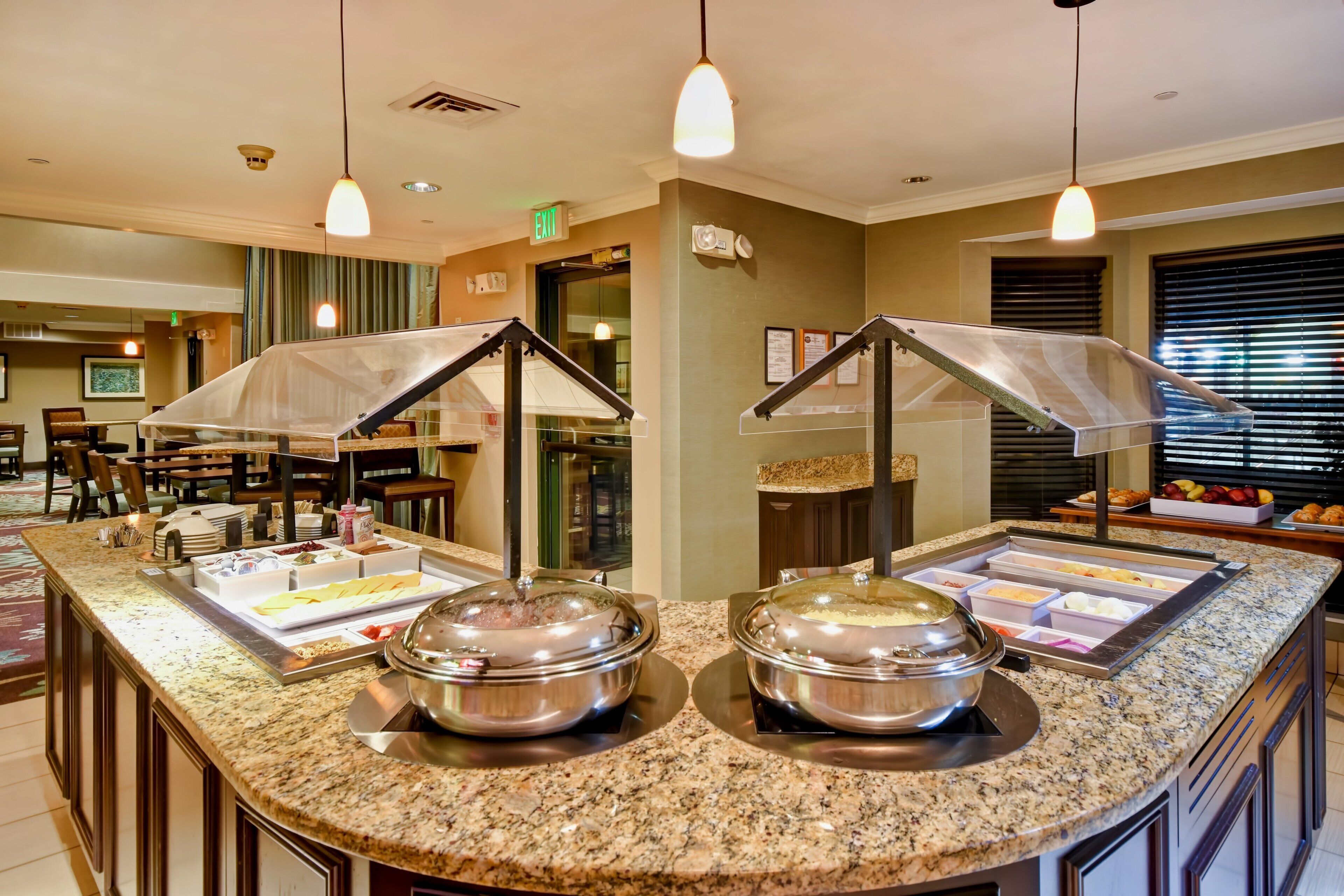 A buffet area with two self-service stations, a seating area with tables and chairs, and a window with blinds.