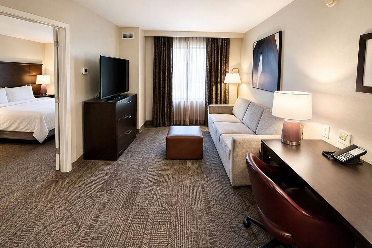 A hotel room with a bed, a TV, a desk with a lamp, a sofa, and a chair.