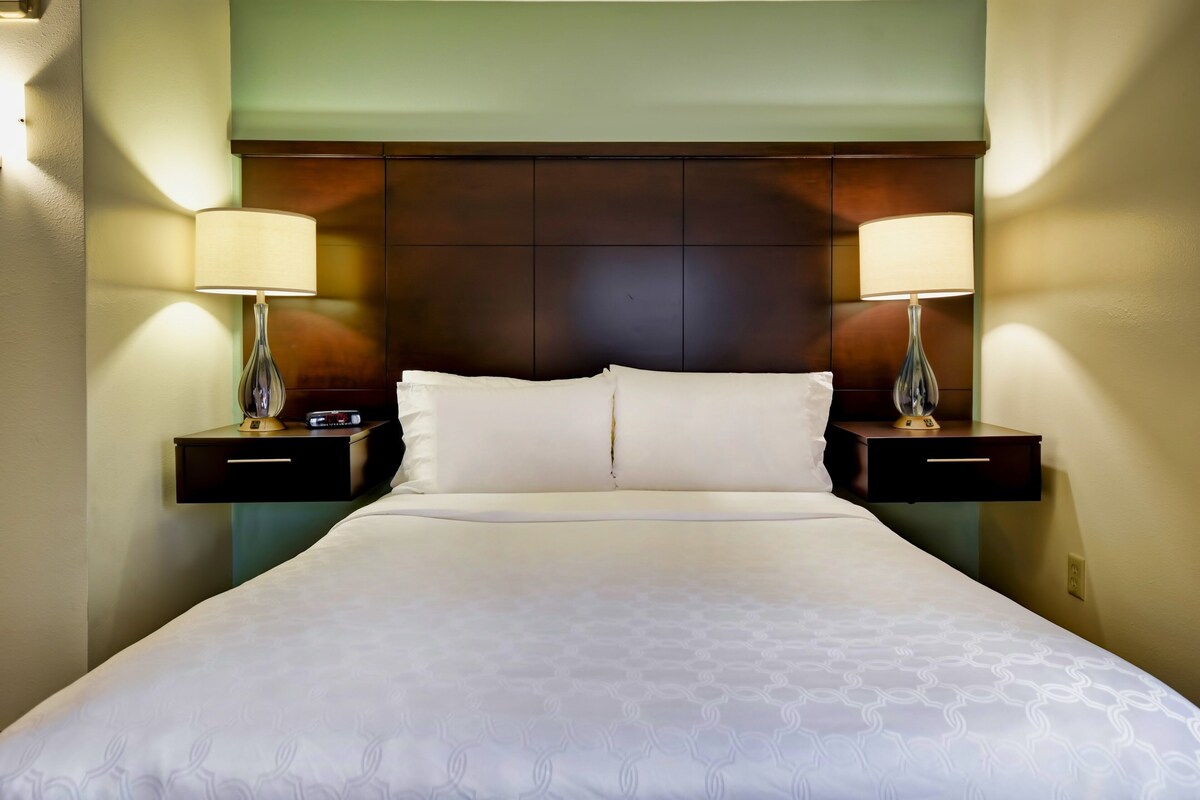 A hotel room with a large bed, two bedside tables with lamps, a headboard, and a wall-mounted nightstand.