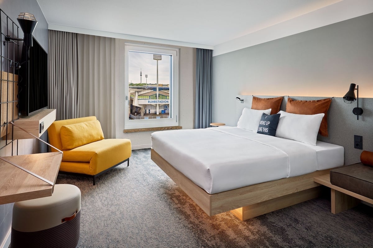 A modern hotel room with a large bed, a yellow armchair, and a view of a stadium.
