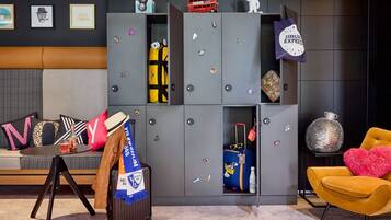 Ein modernes Hotelzimmer mit einem grauen Kleiderschrank, einer Couch mit bunten Kissen, einem kleinen Tisch mit einem Getränk, einem Koffer und einem leuchtend gelben Sessel.