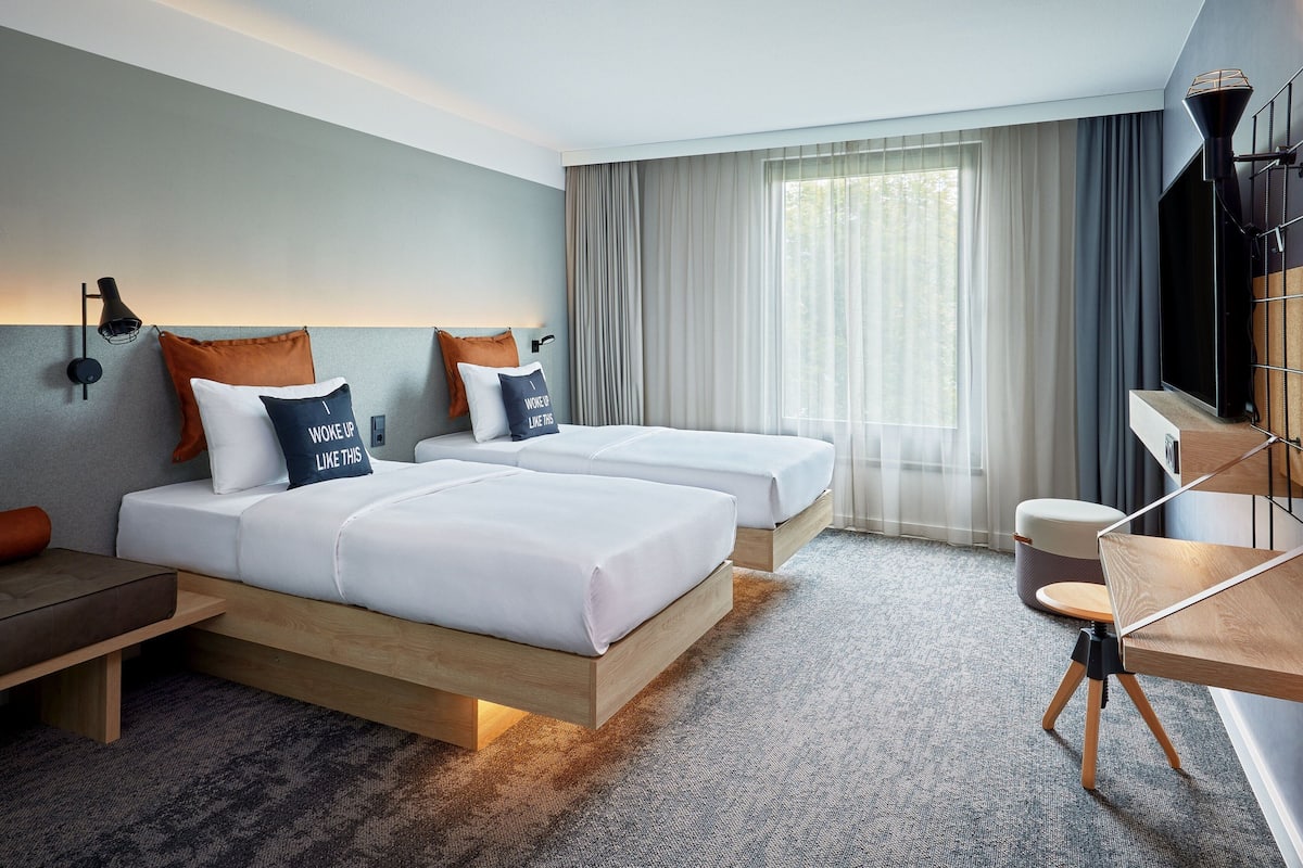 A hotel room with two beds, a desk, a chair, a lamp, and a large window with curtains.
