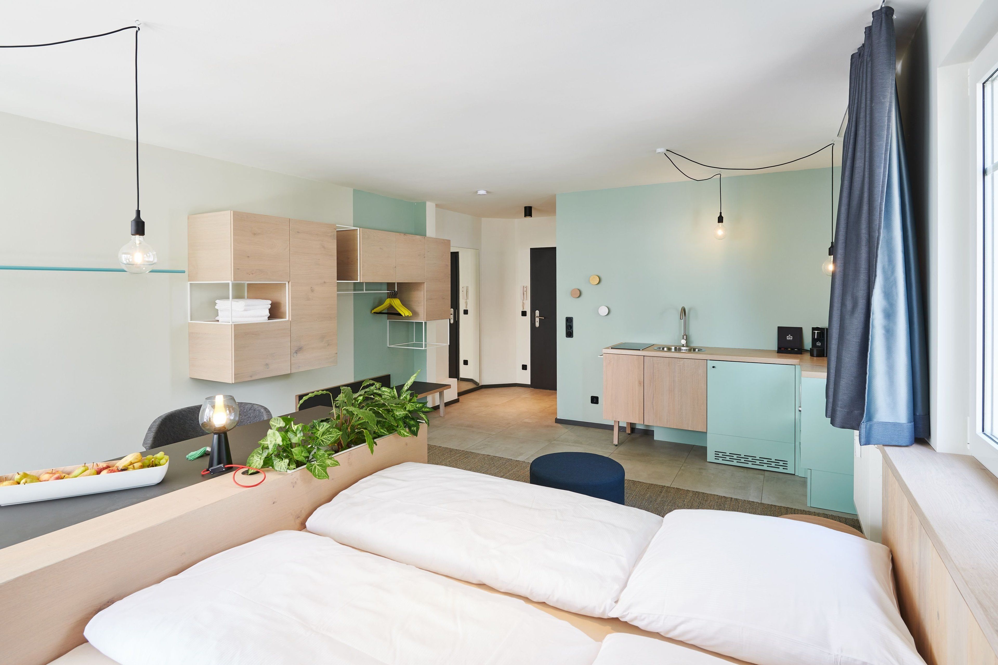 A modern hotel room with a bed, a small desk, a kitchenette, and a bathroom.