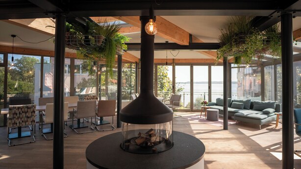 A modern lounge area with a central fire feature, seating arrangements, and large windows offering views of the outdoors. - Hotel Whitman (Ascheberg)
