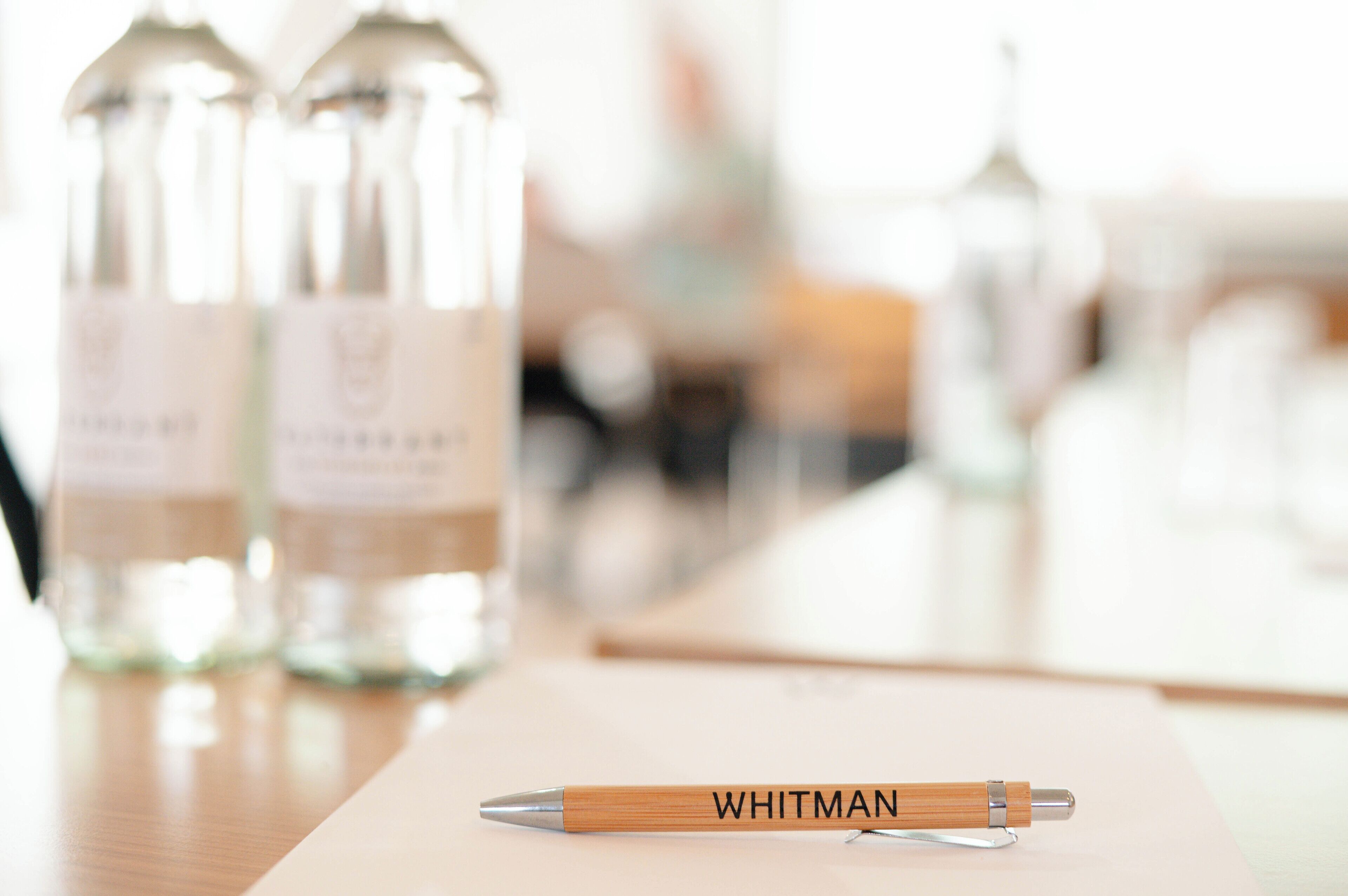 Een houten pen met de merknaam "WHITMAN" op een tafel, met een vage achtergrond van flessen.