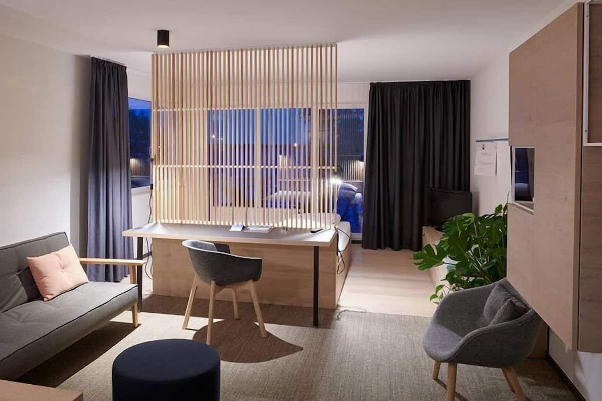 Una sala de estar moderna con un sofá, un escritorio y una silla. Hay un ventanal con cortinas, una planta y vistas al exterior.