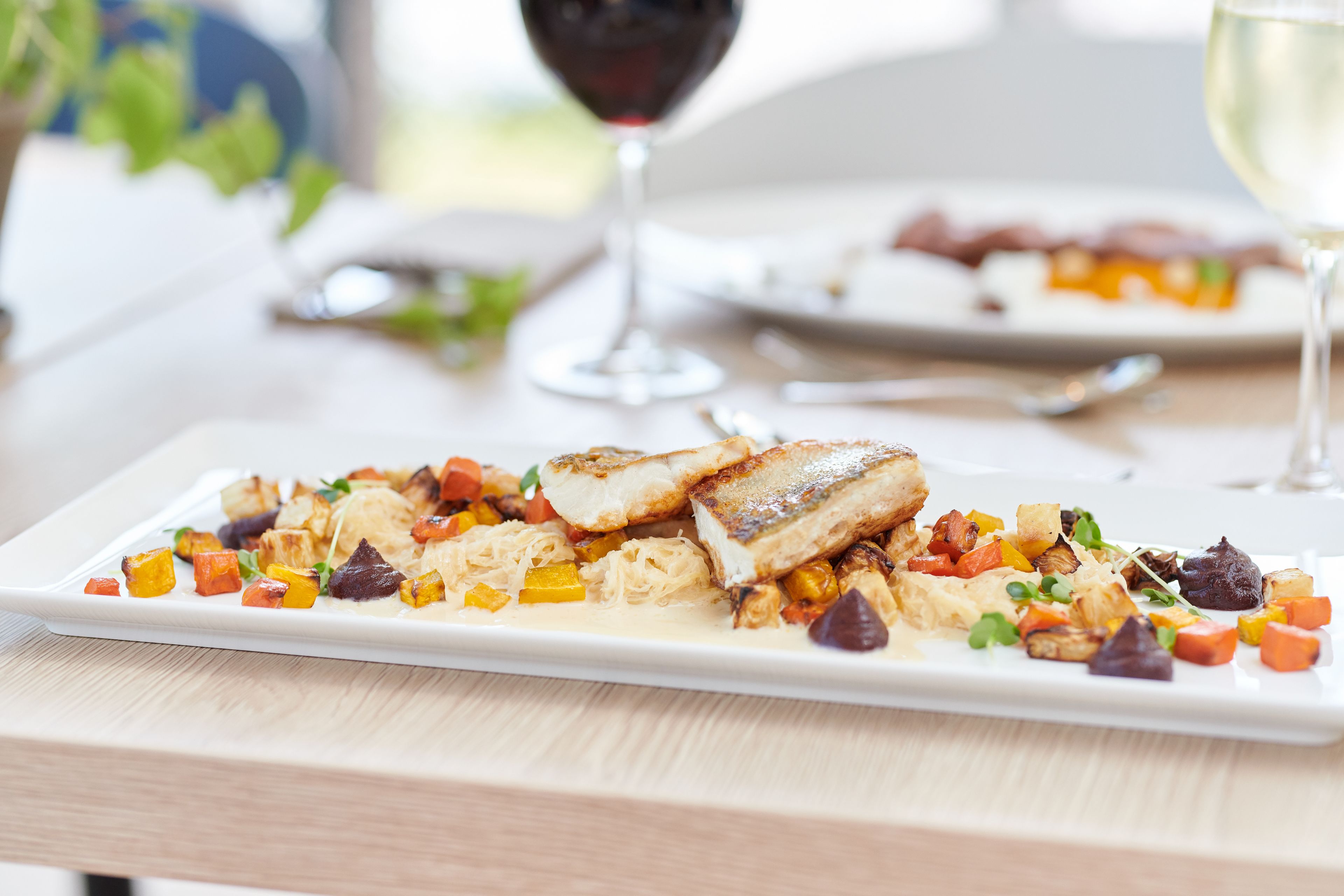 Un plato rectangular blanco con un trozo de pescado a la parrilla, verduras asadas y una salsa cremosa.