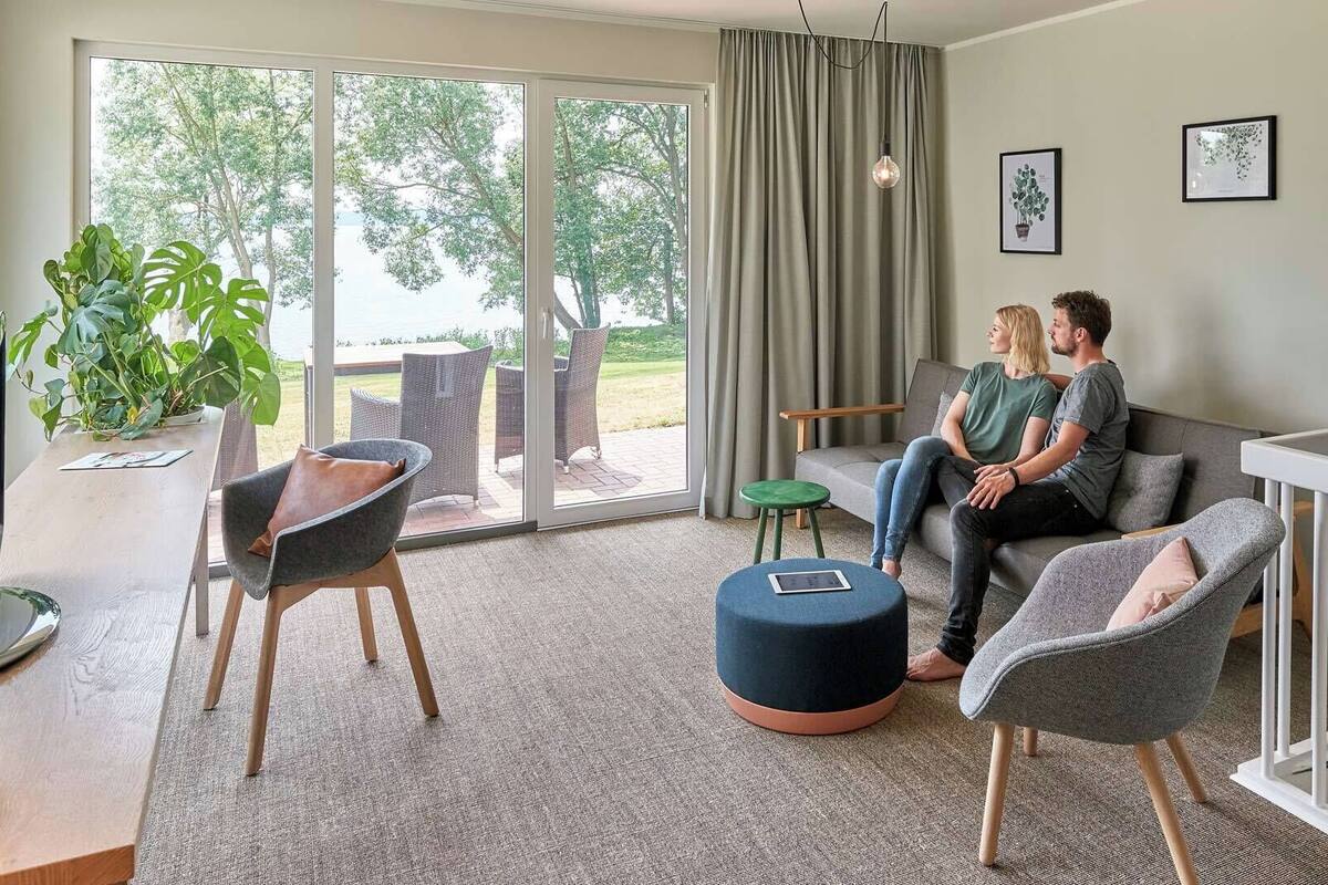Una sala de estar moderna con un sofá, sillas, una mesa de centro y un televisor. Hay un balcón con vistas a árboles y un cuerpo de agua.