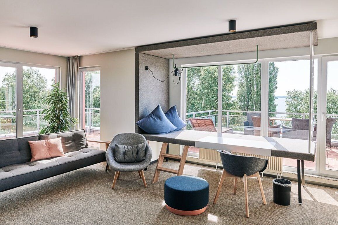 Ein modernes Wohnzimmer mit einer Couch, zwei Sesseln, einem Esstisch und großen Fenstern mit Blick auf Grünflächen und ein Gewässer.