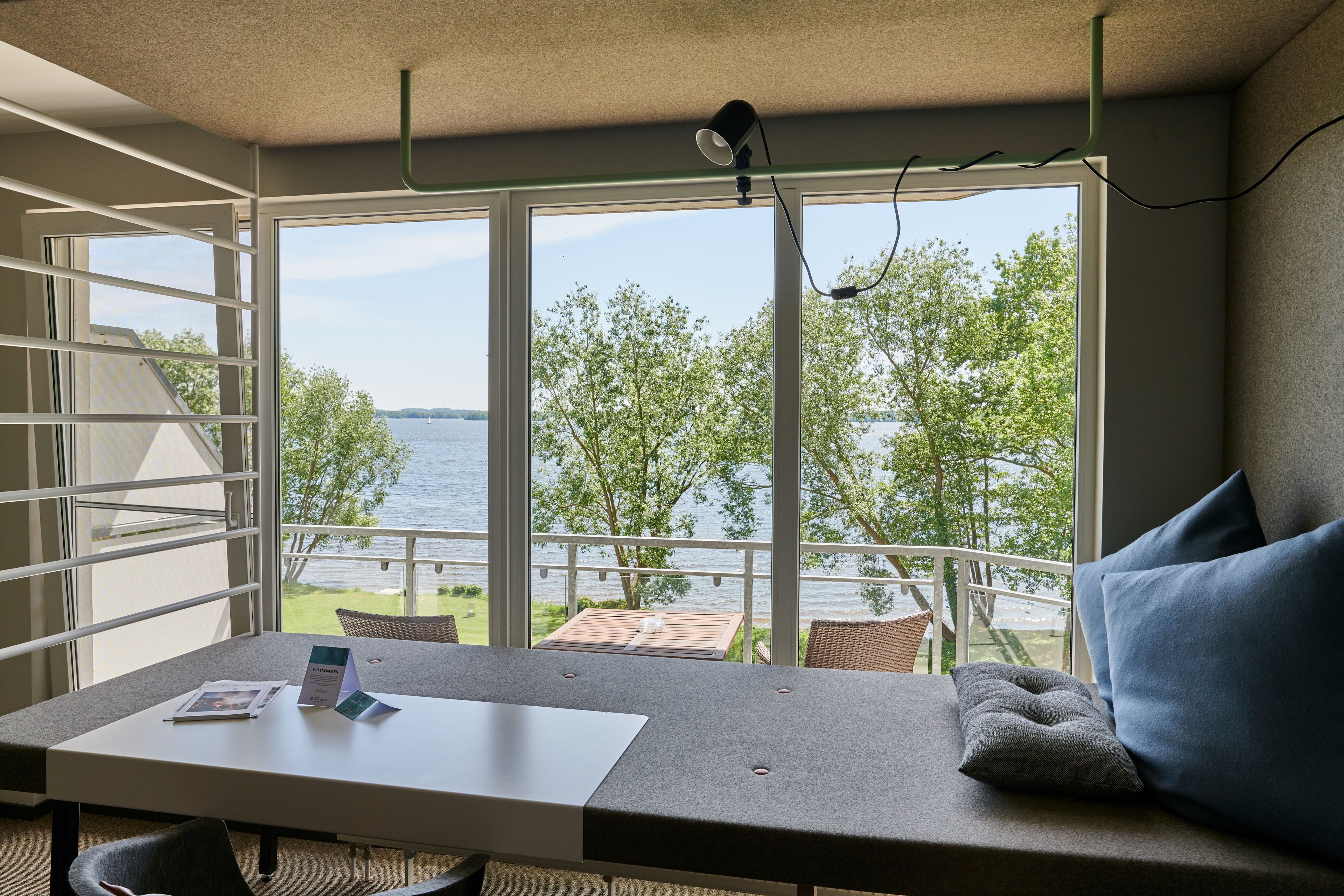 A room with a large window overlooking a body of water, a table with a chair, and a sofa with cushions.