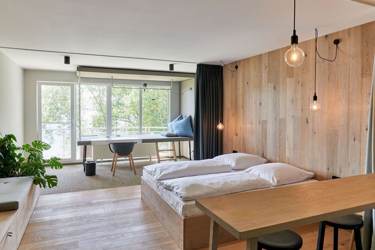 Una habitación moderna con una pared de madera, una cama con ropa de cama blanca, un escritorio con una silla y un ventanal con vistas a árboles.