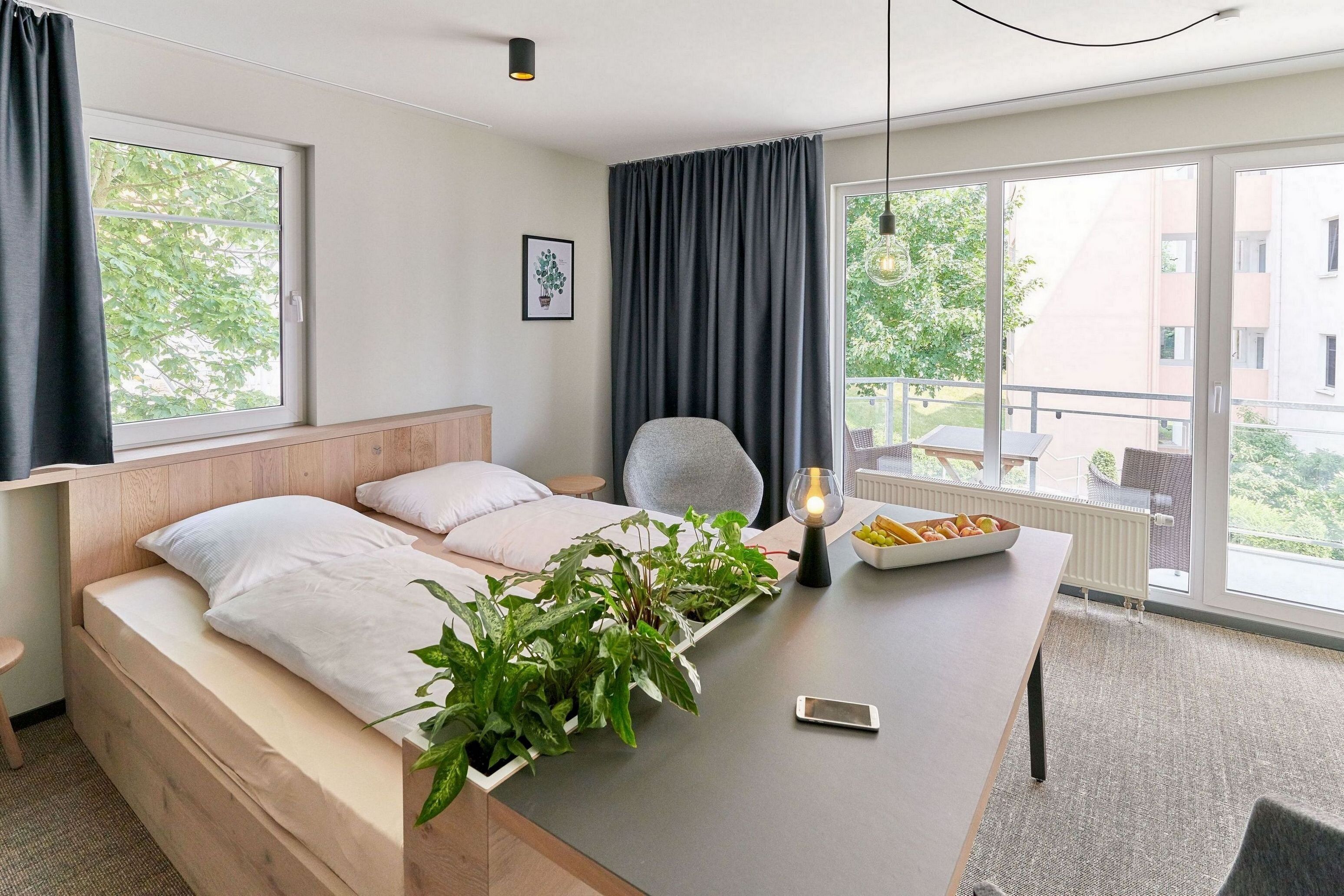 A modern hotel room with a bed, a desk with a lamp, a chair, and a view of the outside.