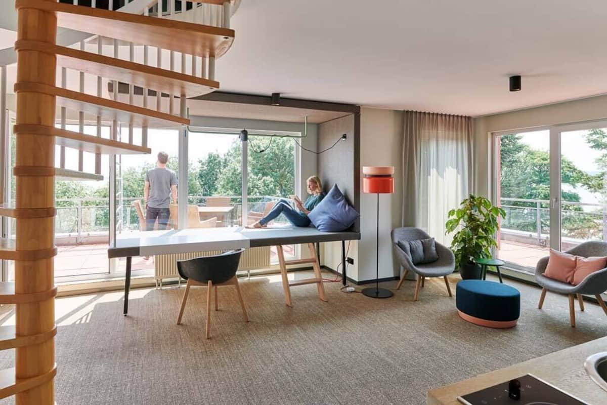 Una sala de estar moderna con una escalera, un comedor y ventanas amplias que ofrecen vistas al exterior.