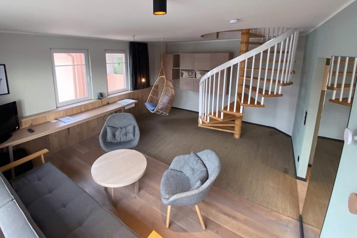 Una sala de estar moderna con un sofá, dos sillones, una mesa de centro, un televisor y una silla colgante. Se observa una escalera con barandilla de madera que conduce a un nivel superior.