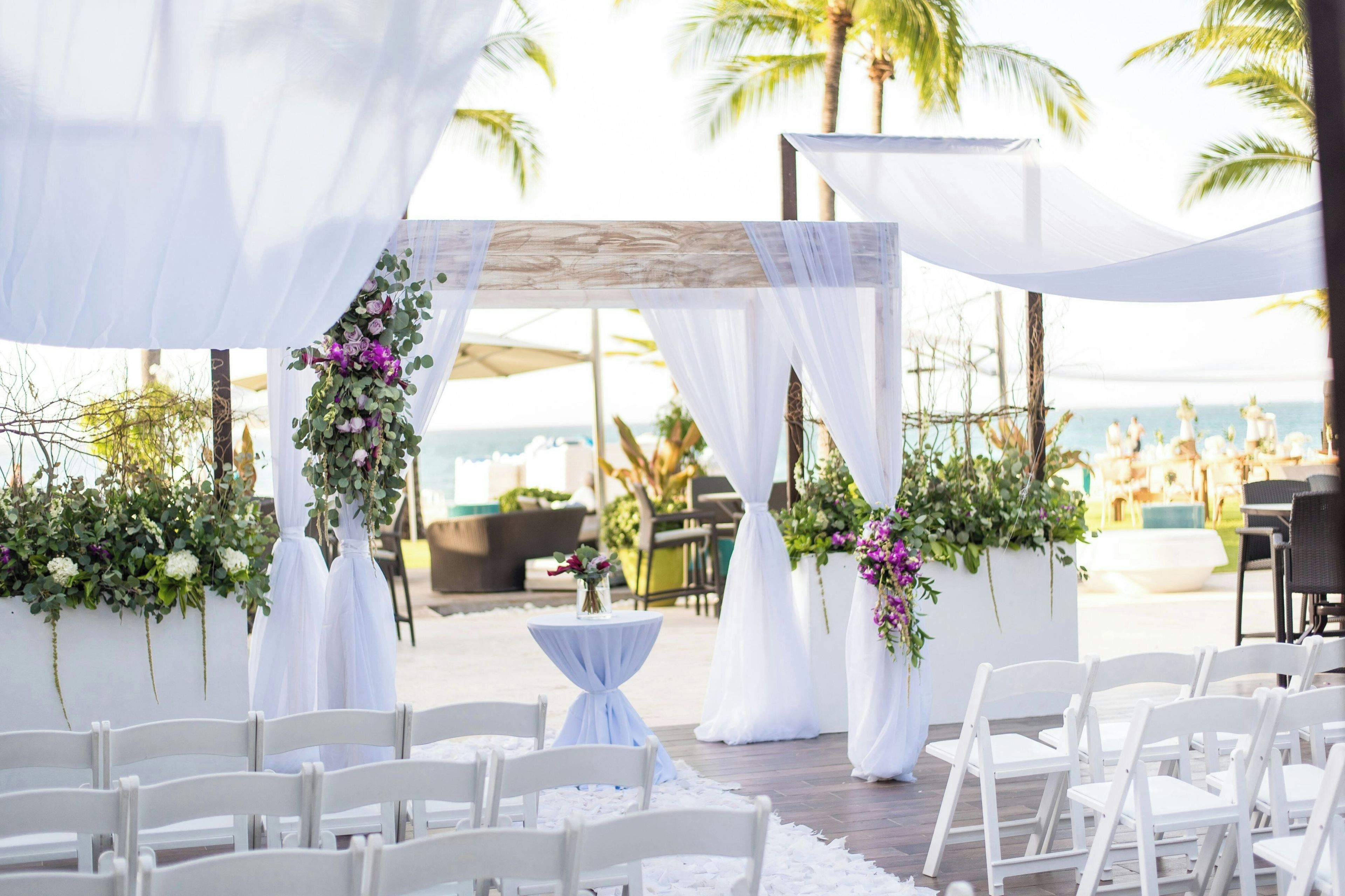 An outdoor wedding venue with white chairs arranged in rows, a small table with a white cloth, and floral decorations.