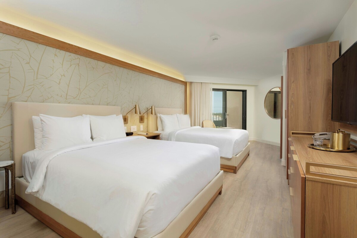 A hotel room with two beds, a desk with a telephone and a small table, a mirror, and a view of the outdoors.