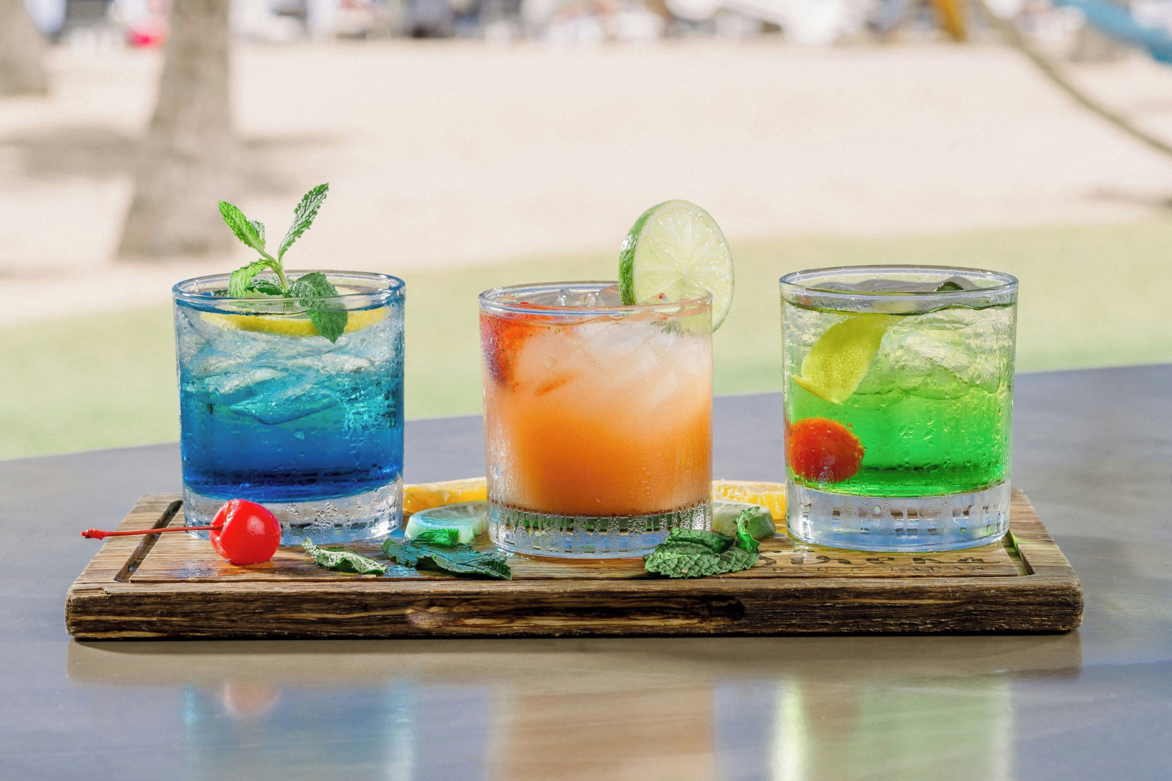 Three glasses of different colored beverages on a wooden table with a cherry and mint leaves.