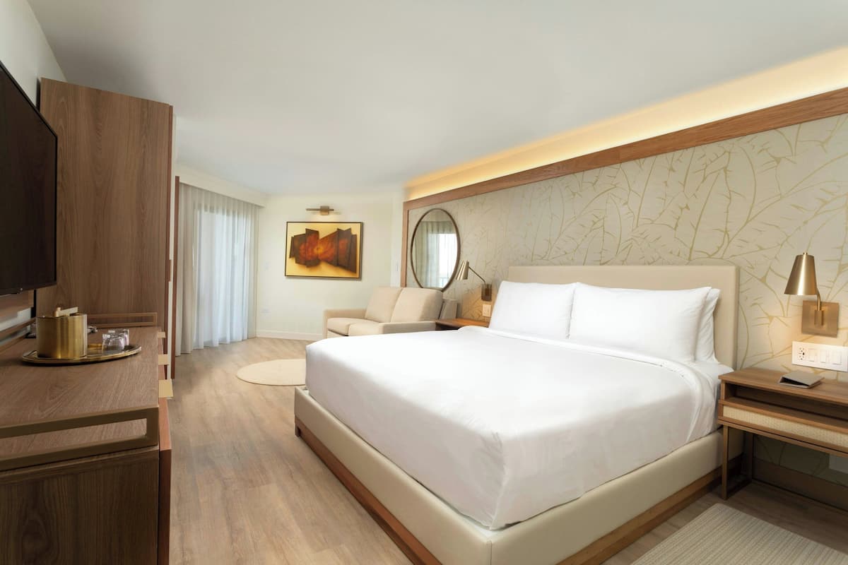 A modern hotel room with a large bed, a desk with a lamp, a chair, and a television.
