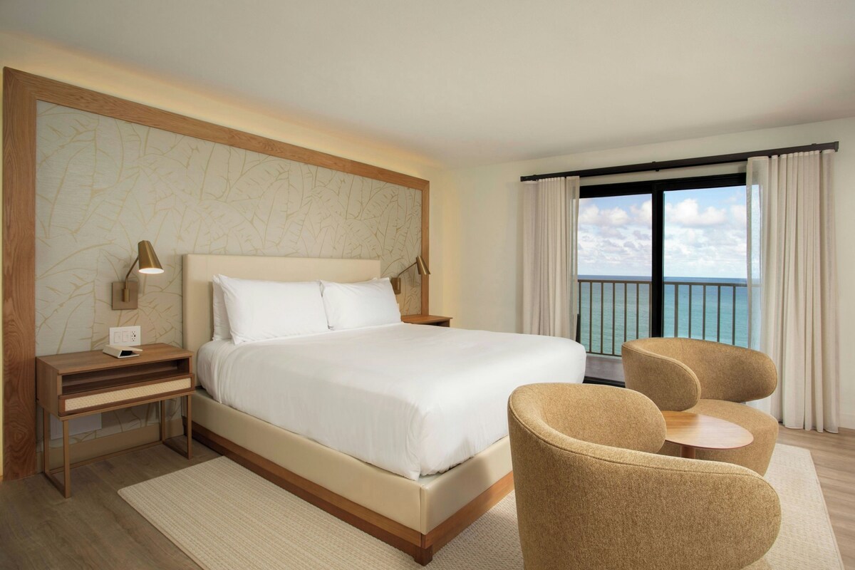 Una habitación de hotel con una cama grande, mesitas de noche, un escritorio, dos sillones, vista al mar por la ventana y un papel tapiz estampado.