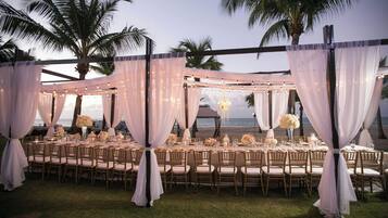 Montagem de banquete ao ar livre, com mesas e cadeiras cobertas por tecido branco e luzes de corda.