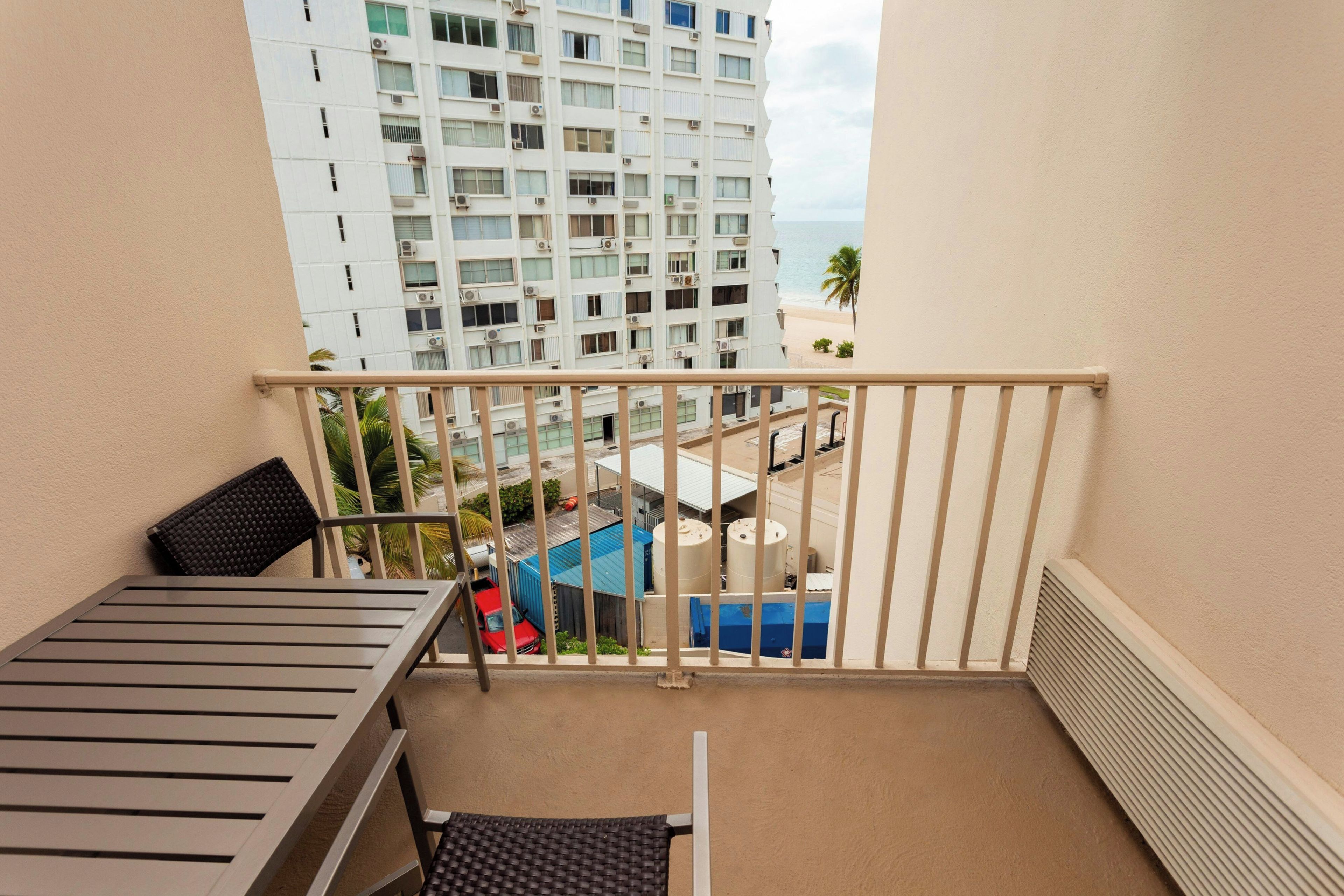A balcony with a view of a tall building, a red vehicle, and some industrial structures.