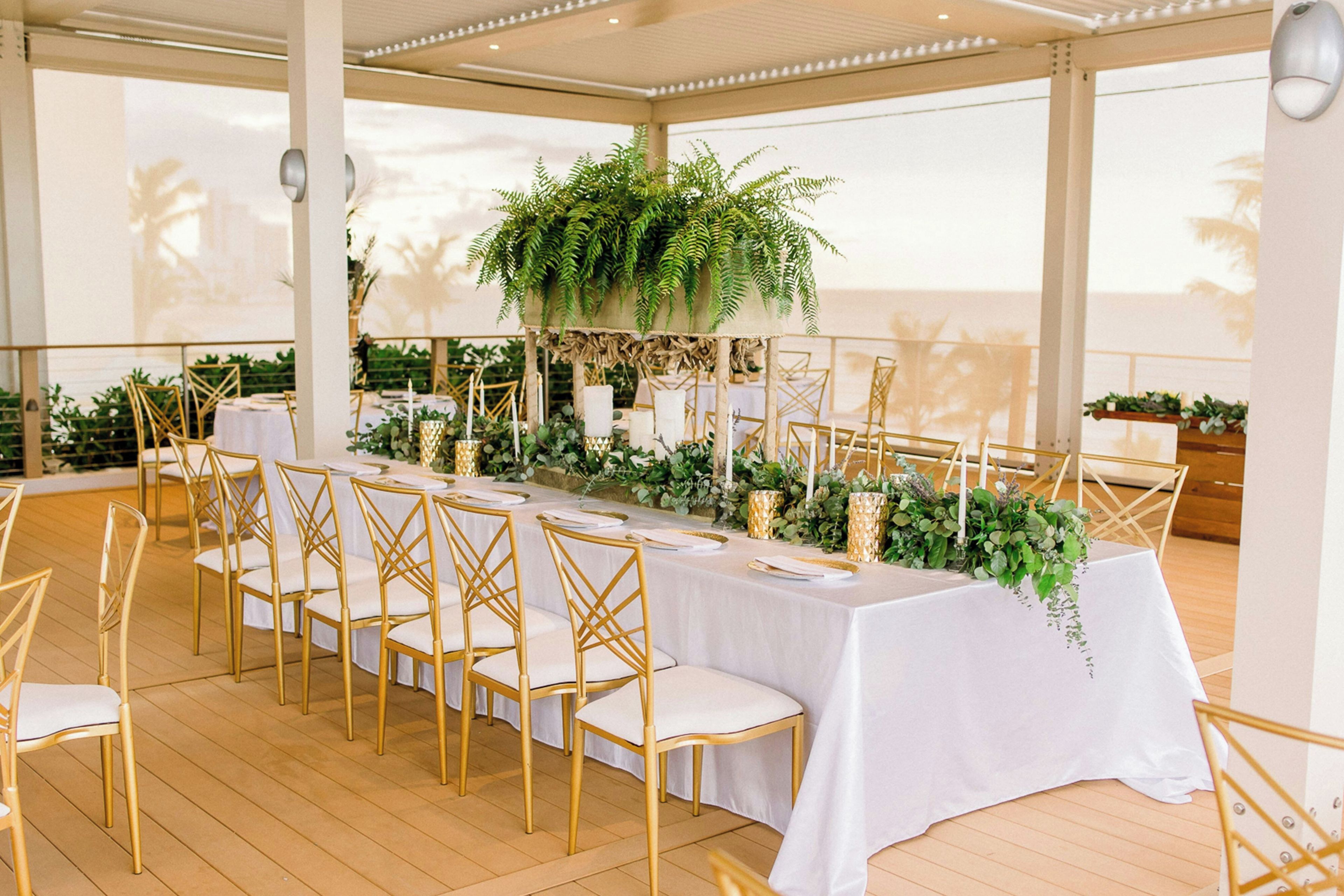 A long table set for a formal event with gold chairs, white tablecloth, and greenery decorations.