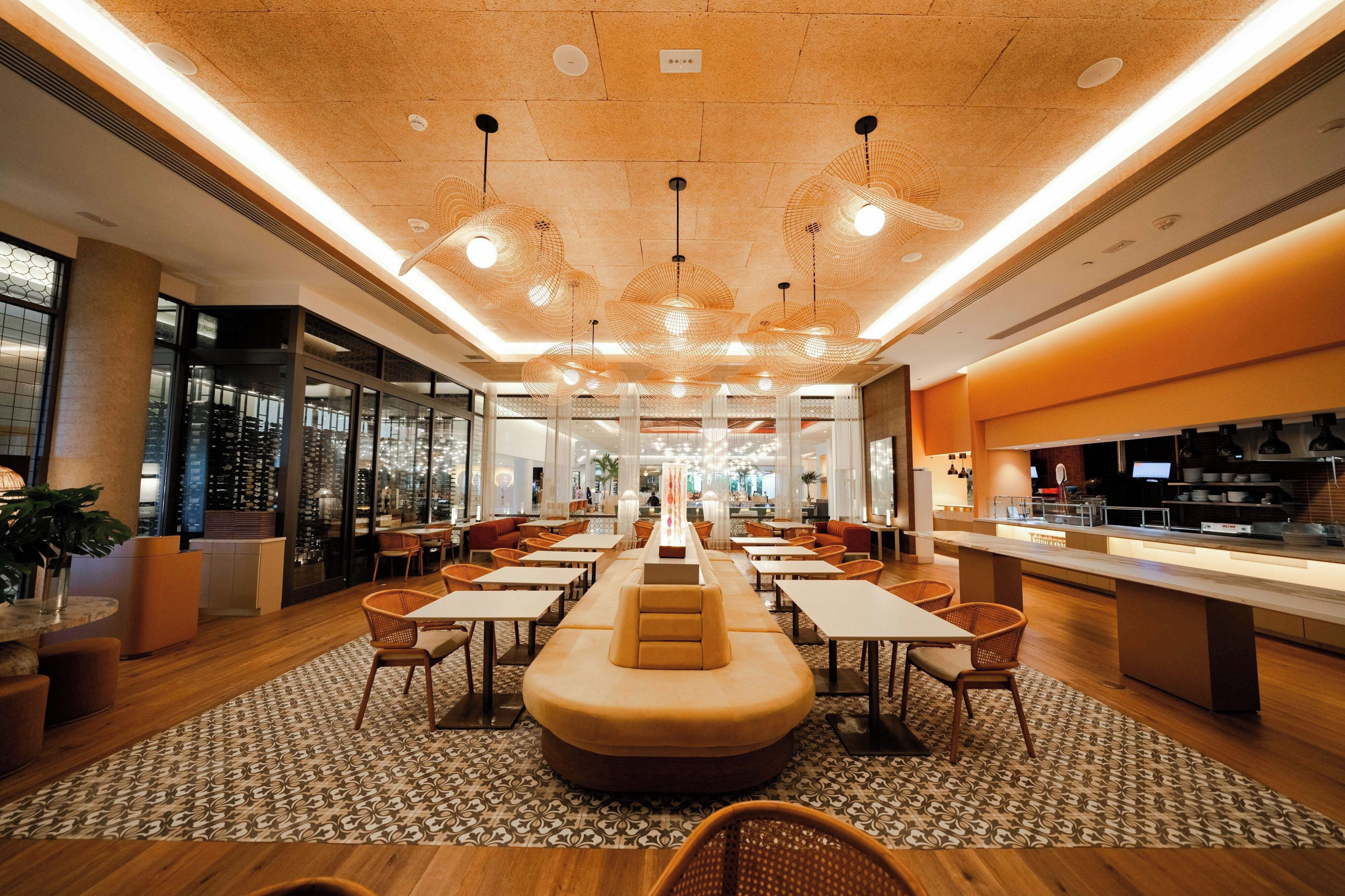 A modern hotel lobby with a central seating area, multiple tables, and a kitchen visible to the right.