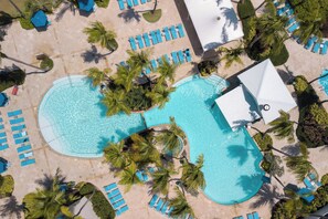 Luchtbeeld van een resort met een groot zwembad, omringd door palmbomen en blauwe ligbedden.