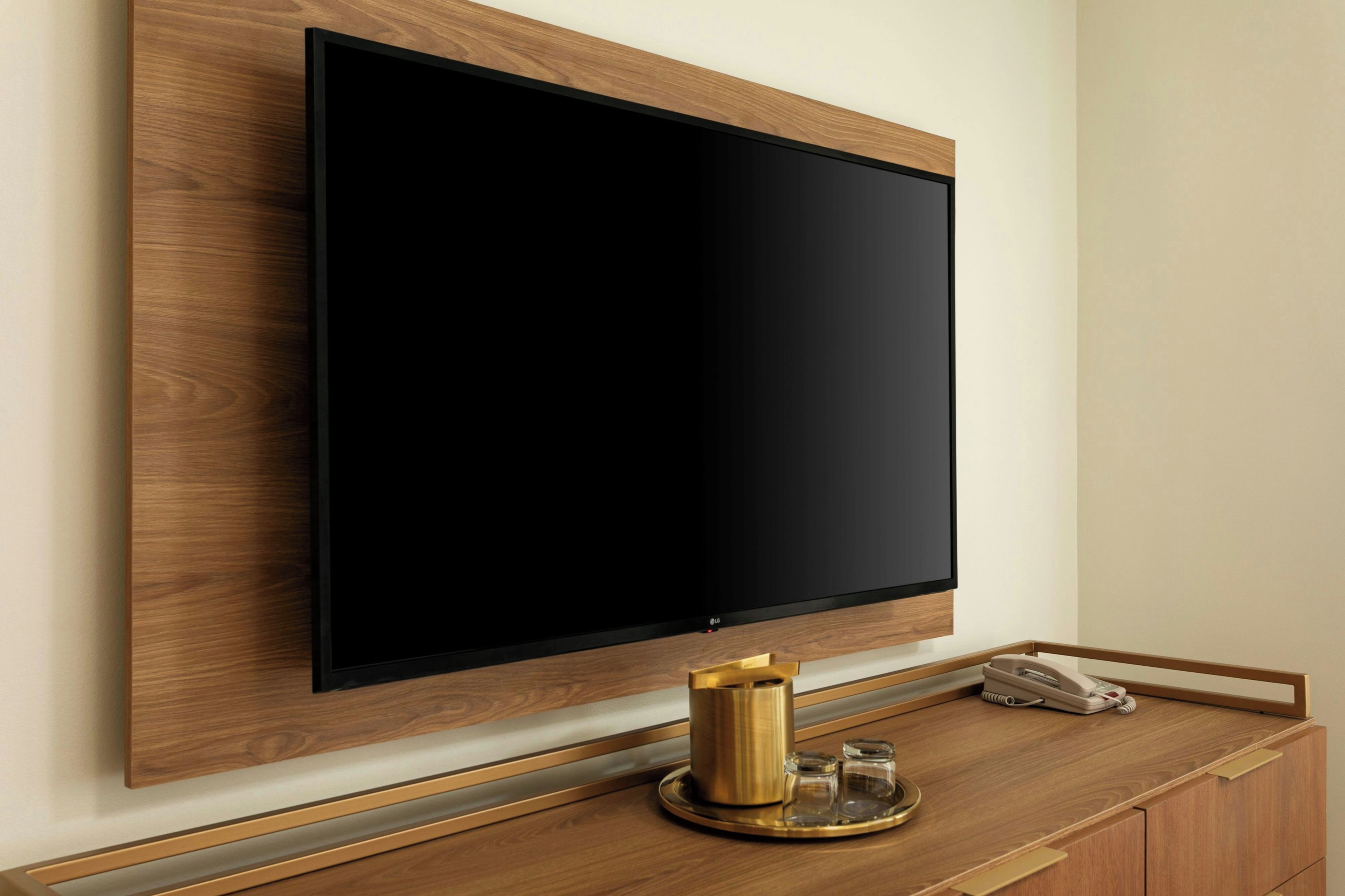 A flat-screen TV mounted on a wooden wall, a brass coffee pot, and a telephone on a wooden counter.