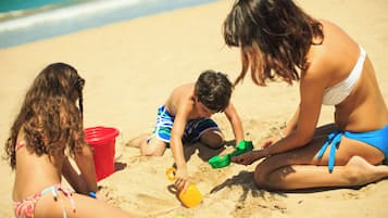 Three individuals on a sandy beach with a red bucket and green and yellow sand toys.