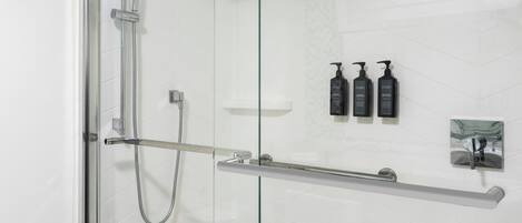 A modern bathroom with a glass shower enclosure, a handheld showerhead, a glass shelf with toiletries, and a towel rack.