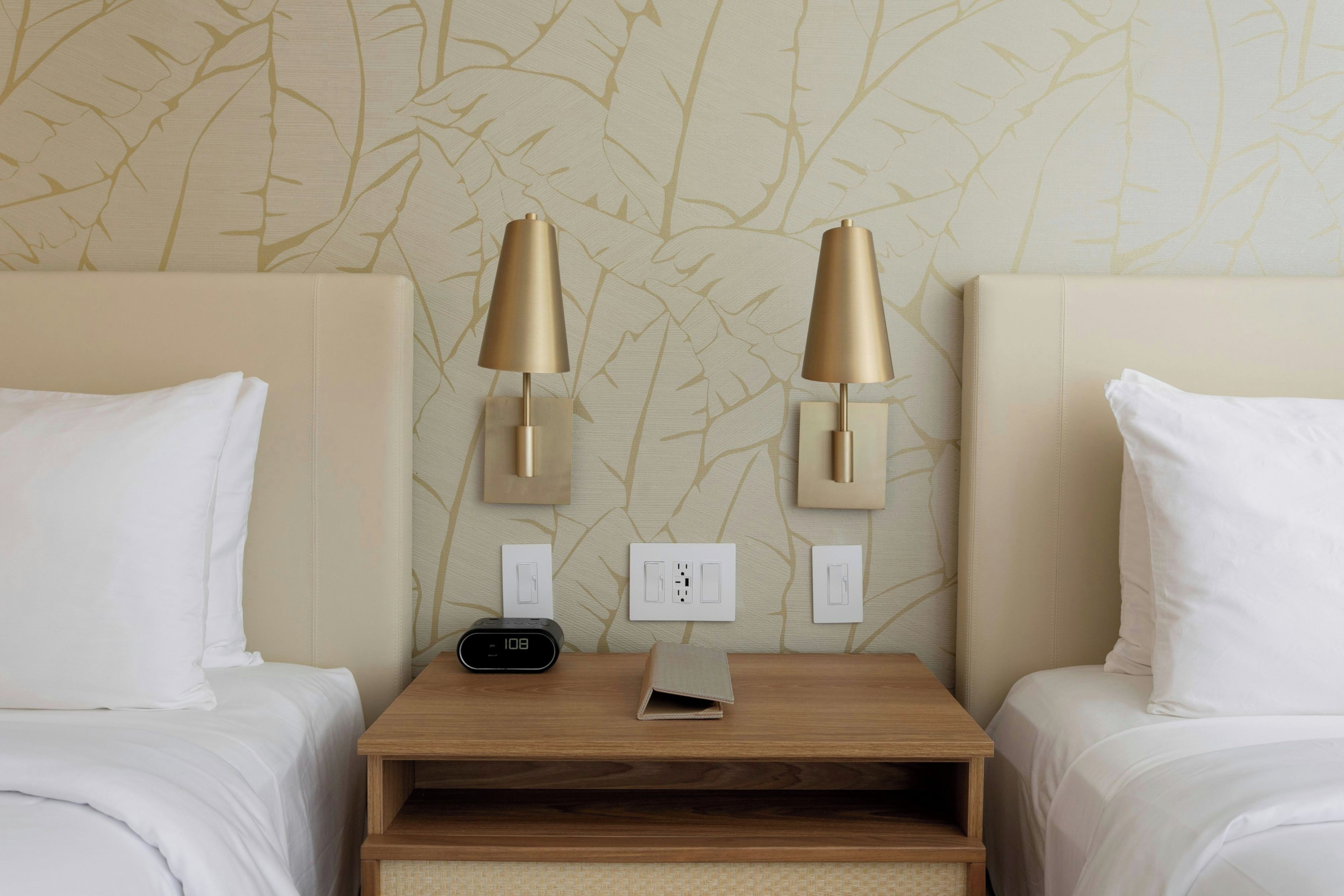 A hotel room with two beds, a nightstand, a digital clock, and wall-mounted lamps.