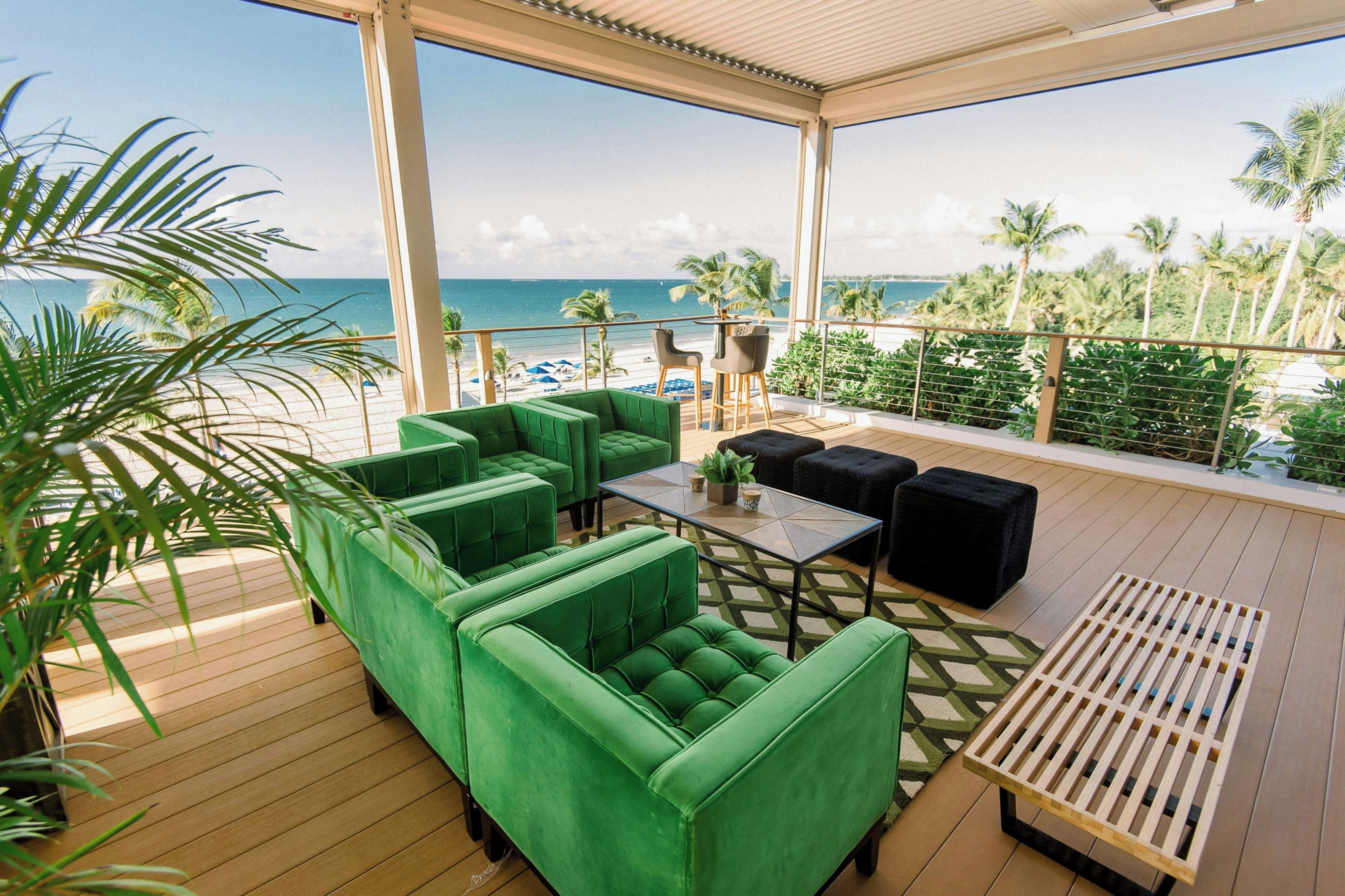 Un espacio de descanso al aire libre con sofás tapizados en tonos verdes y una mesa, con vistas a una playa con palmeras y el océano.