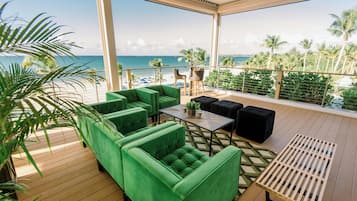 Un espacio de descanso al aire libre con sofás tapizados en tonos verdes y una mesa, con vistas a una playa con palmeras y el océano.