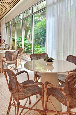 A spacious indoor area with a round table surrounded by wicker chairs, large windows with curtains, and a view of lush greenery outside.