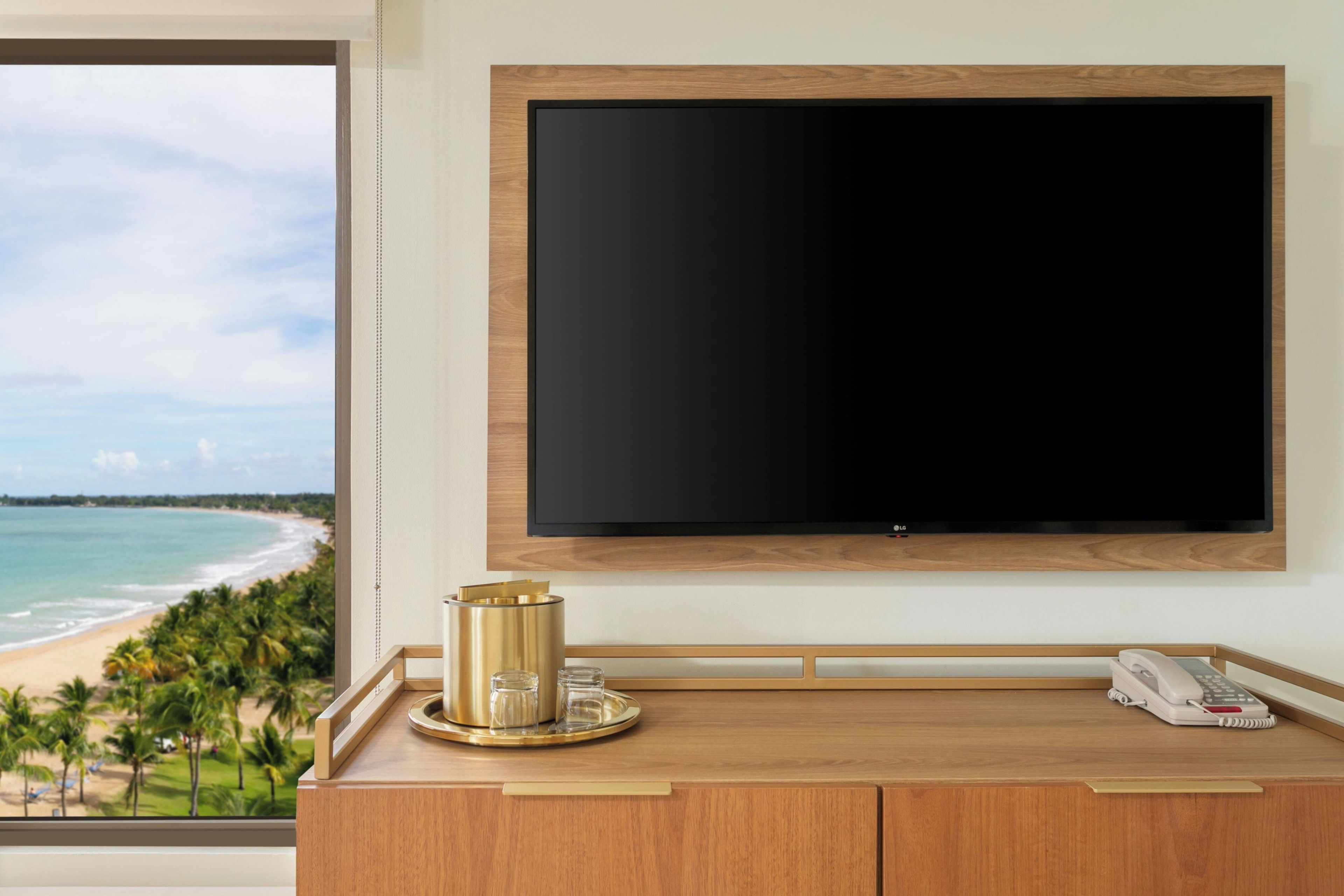 Ett hotellrum med en stor, platt-TV som är monterad på väggen, ett träbord med en telefon och en dekorativ prydnad, och utsikt över en strand genom ett stort fönster.