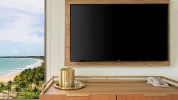Ett hotellrum med en stor, platt-TV som är monterad på väggen, ett träbord med en telefon och en dekorativ prydnad, och utsikt över en strand genom ett stort fönster.