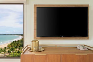 A hotel room with a large flat-screen TV mounted on the wall, a wooden console table with a telephone and a decorative item, and a view of a beach through a large window.
