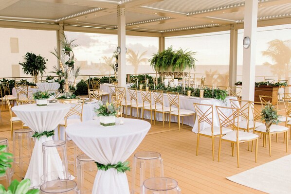 Un espacio para eventos en interior con mesas redondas, sillas doradas y plantas.