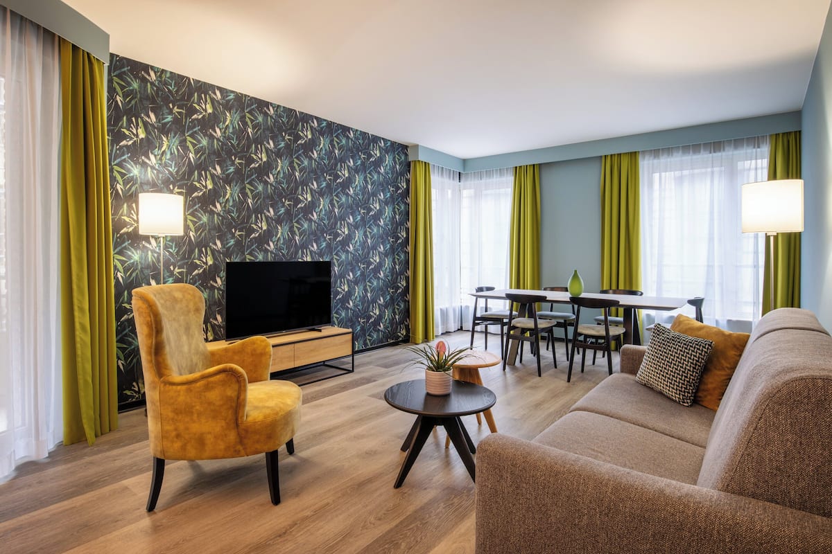A modern living room with a flat-screen TV, a sofa, a yellow armchair, a dining table with chairs, and a green accent wall.