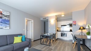 A modern living room with a grey sofa, dining table, and chairs. There is a kitchen in the background with white cabinets and a stove. - Thon Residence Parnasse (Brussels)