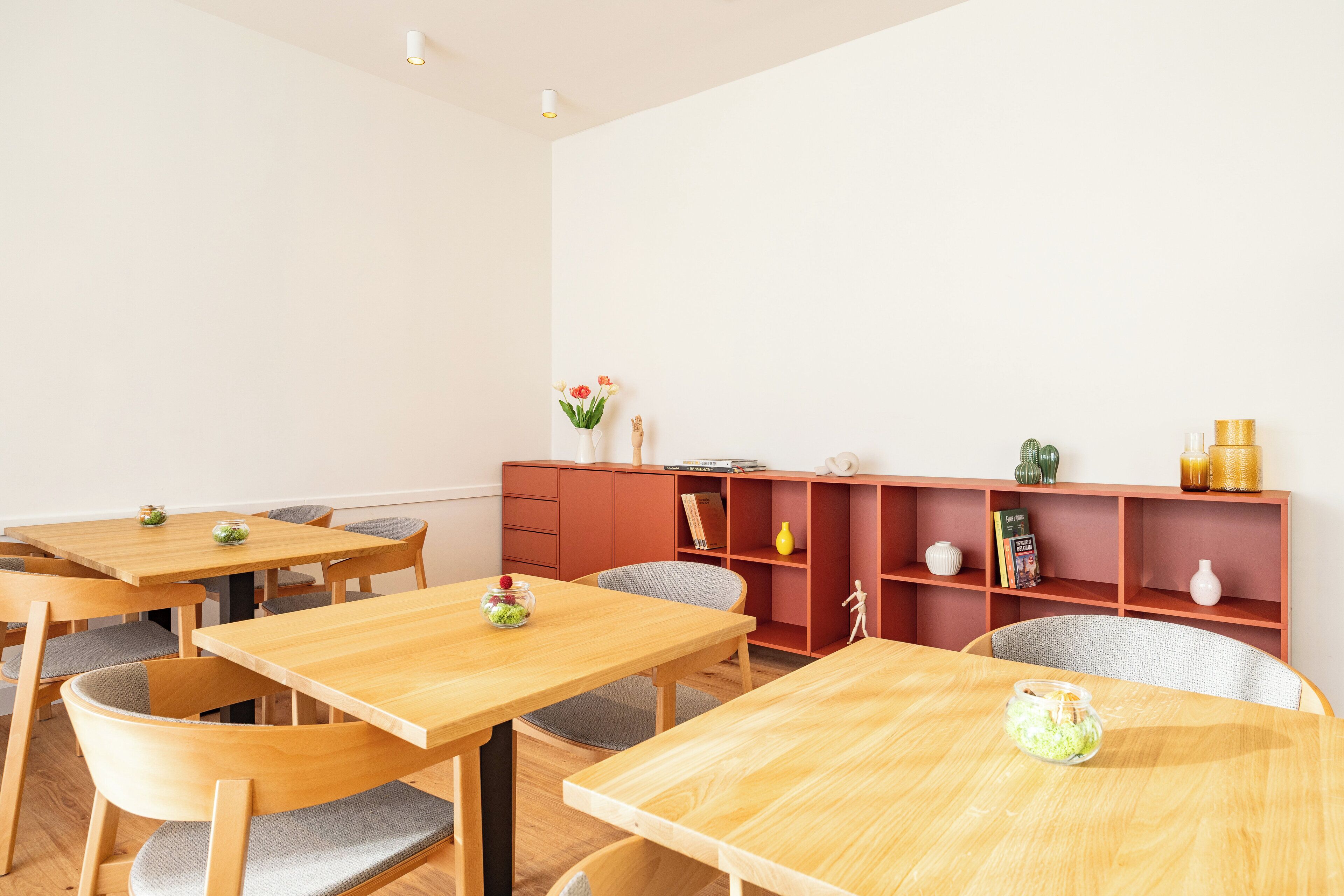 A dining area with wooden tables and chairs, a red bookshelf, and a vase with flowers.