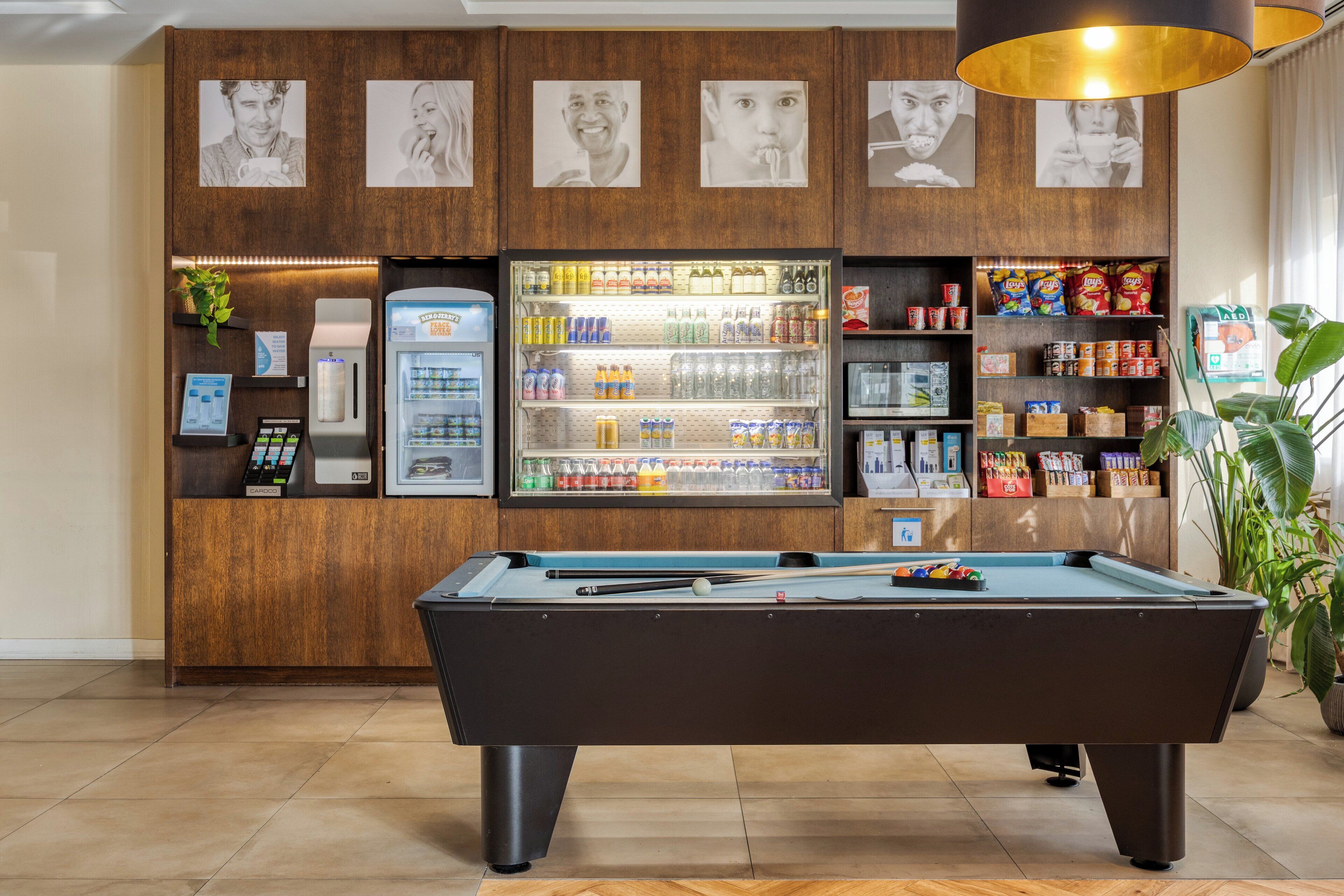 A pool table with a beverage cooler and snack shelves in a hotel lobby.