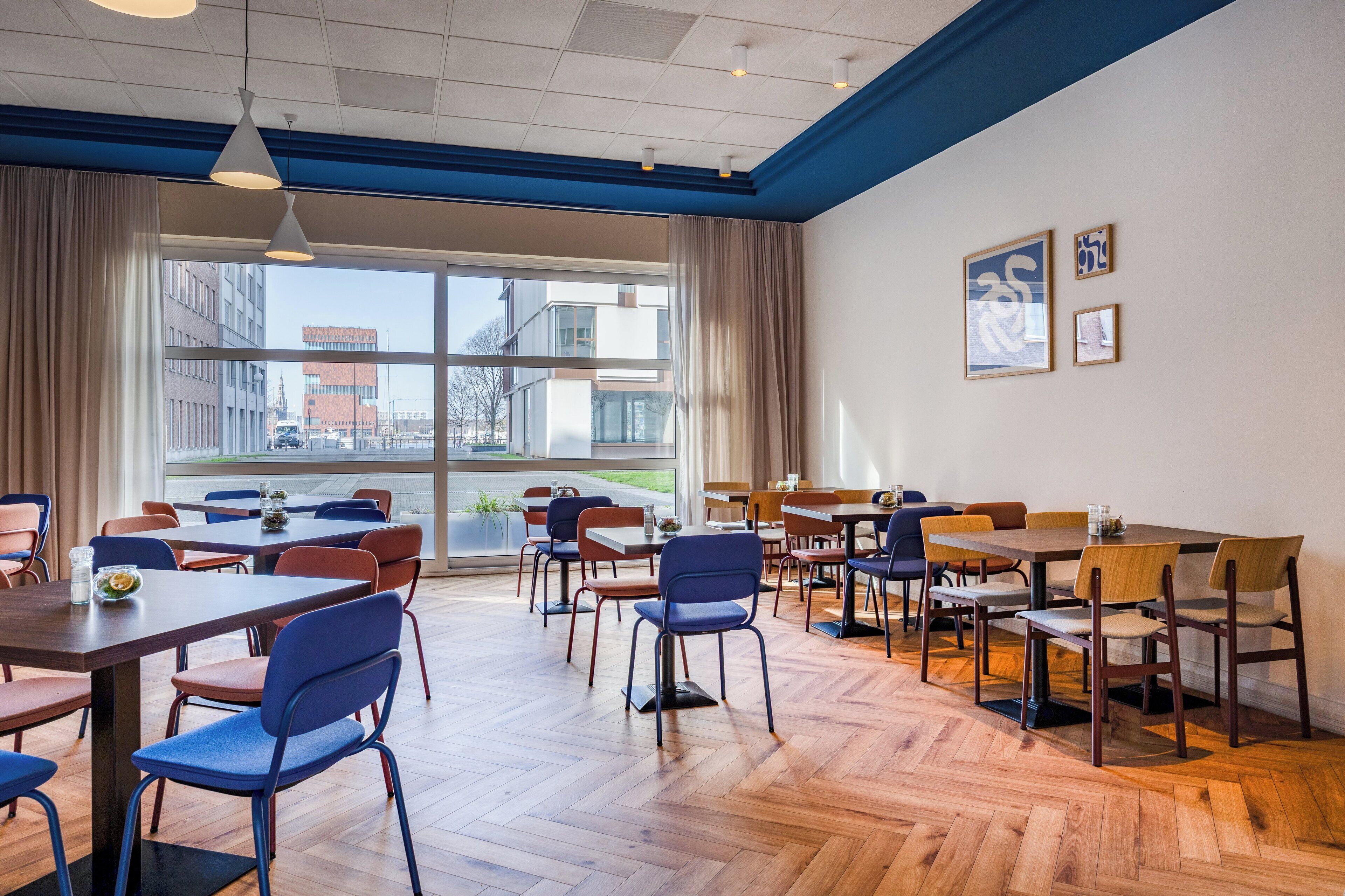 A modern dining area with multiple tables and chairs, large windows with curtains, and framed artwork on the wall.