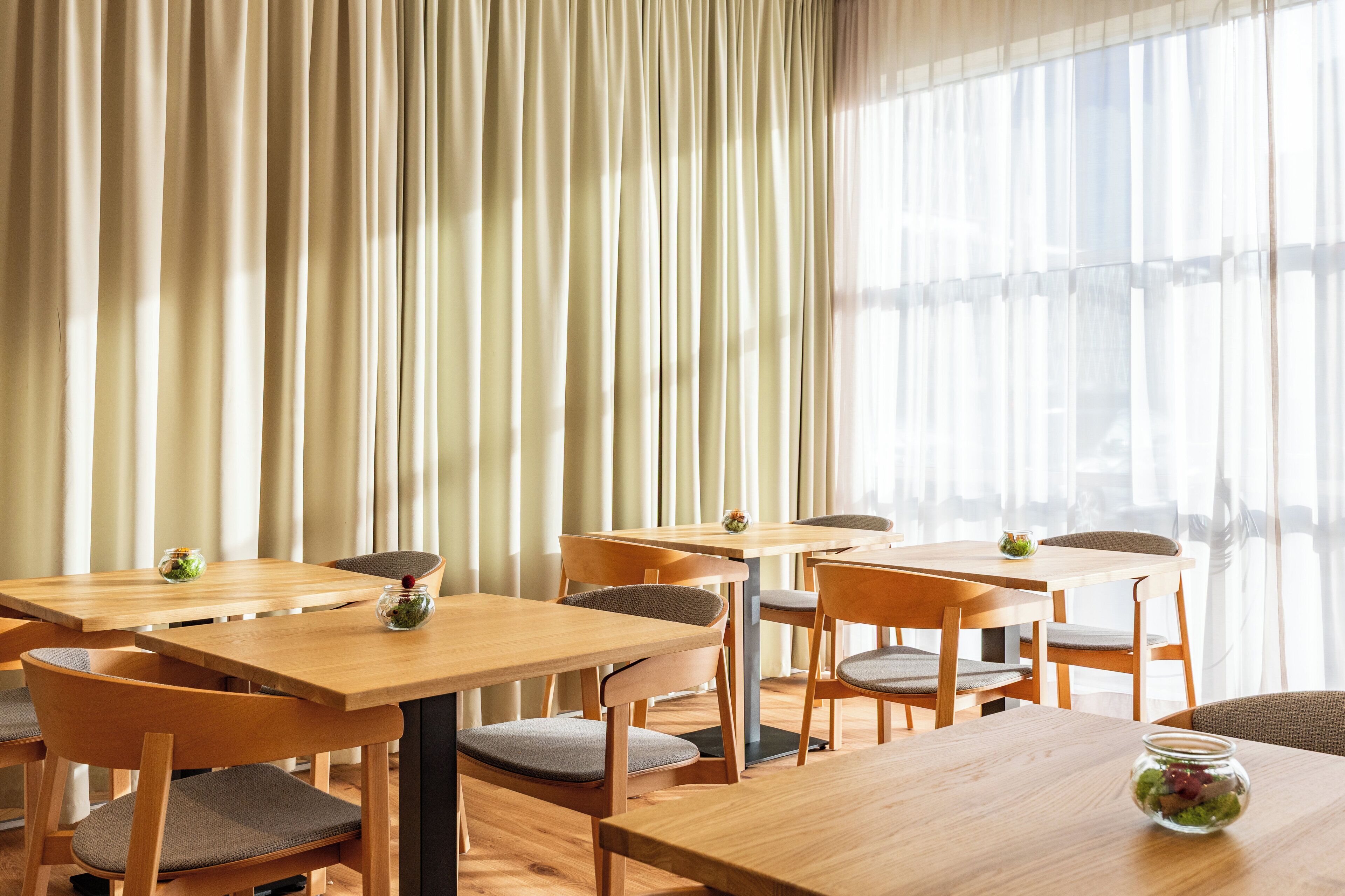 Un espace repas avec des tables et des chaises en bois, de grandes fenêtres ornées de rideaux légers et de petits vases décoratifs posés sur les tables.