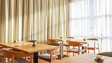 Un espace repas avec des tables et des chaises en bois, de grandes fenêtres ornées de rideaux légers et de petits vases décoratifs posés sur les tables.