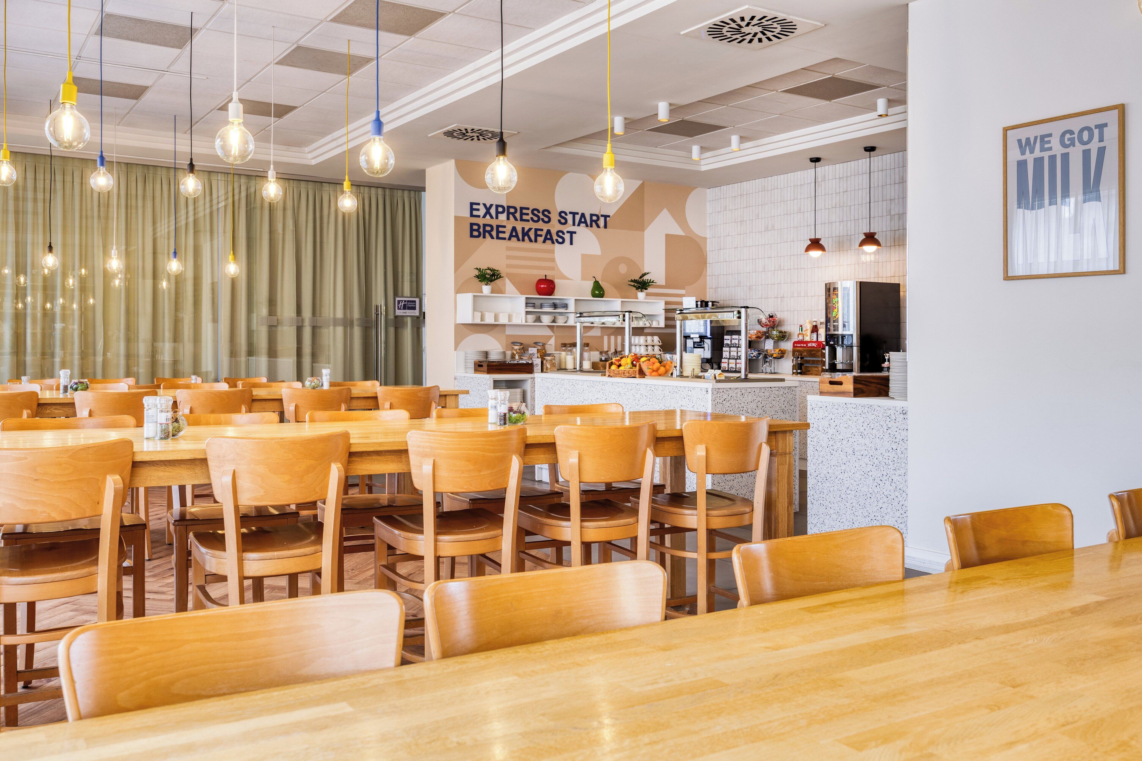 Ein geräumiger Frühstücksbereich mit Holztischen und -stühlen, einer Anrichte mit Speisen und einem Schild mit der Aufschrift 'EXPRESS START BREAKFAST'.