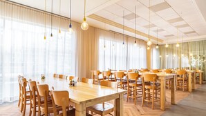 Un vaste espace dédié à la salle à manger, avec de nombreuses tables et chaises en bois, de grandes fenêtres ornées de rideaux légers et des luminaires suspendus.